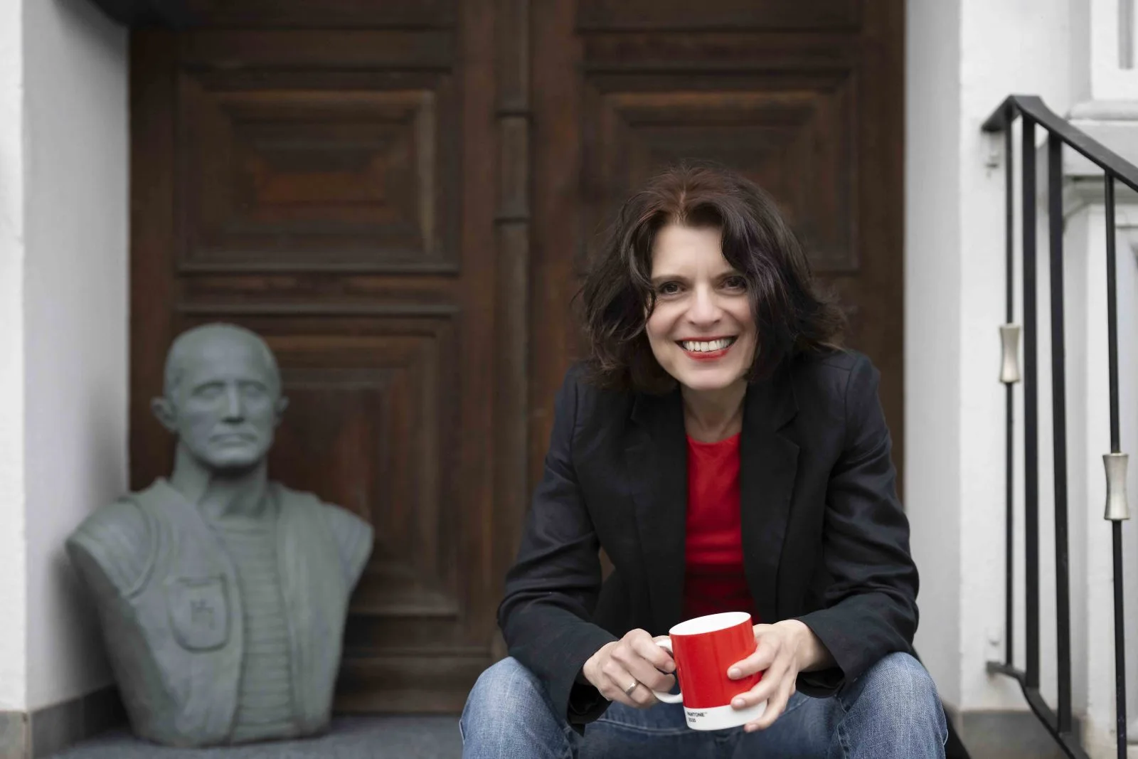 Annette Hardes sitzt mit einer roten Tasse vor einer hölzernen Tür neben einer Steinbüste und lächelt.
