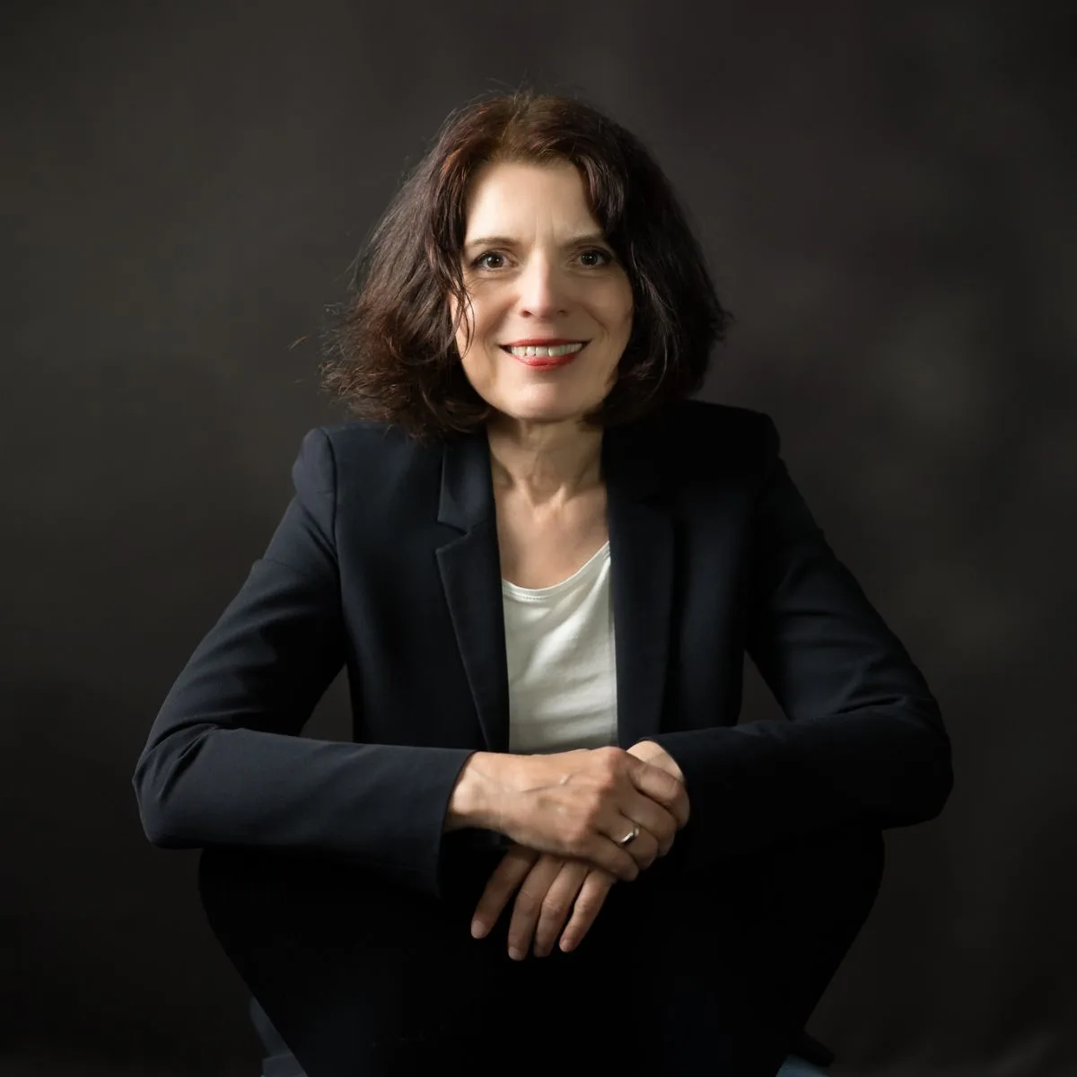 Annette Hardes in dunklem Blazer vor dunklem Hintergrund, sie sitzt leicht vorgebeugt und lächelt in die Kamera.