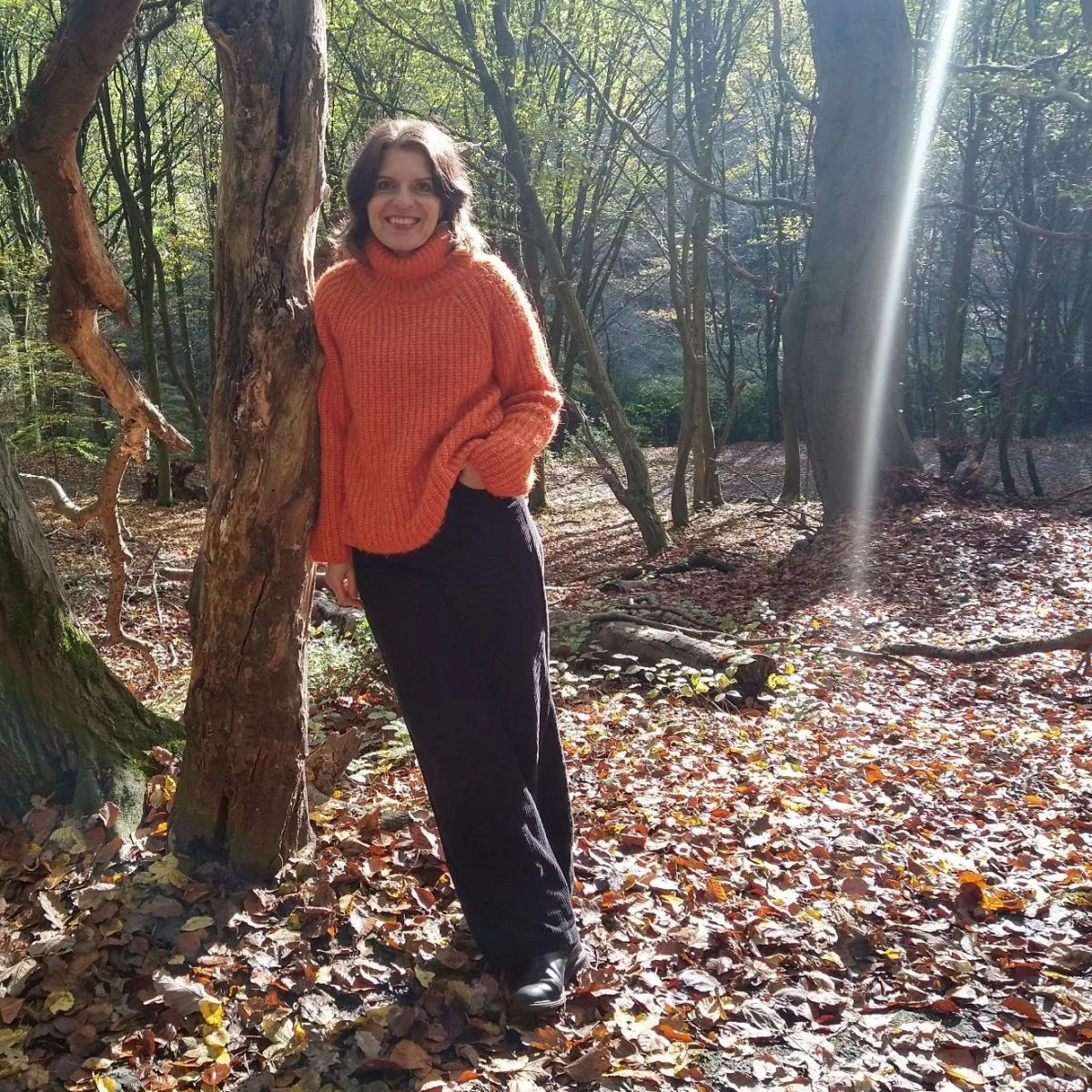 Annette Hardes in rotem Pullover lehnt im herbstlichen Wald an einem Baum und lächelt.